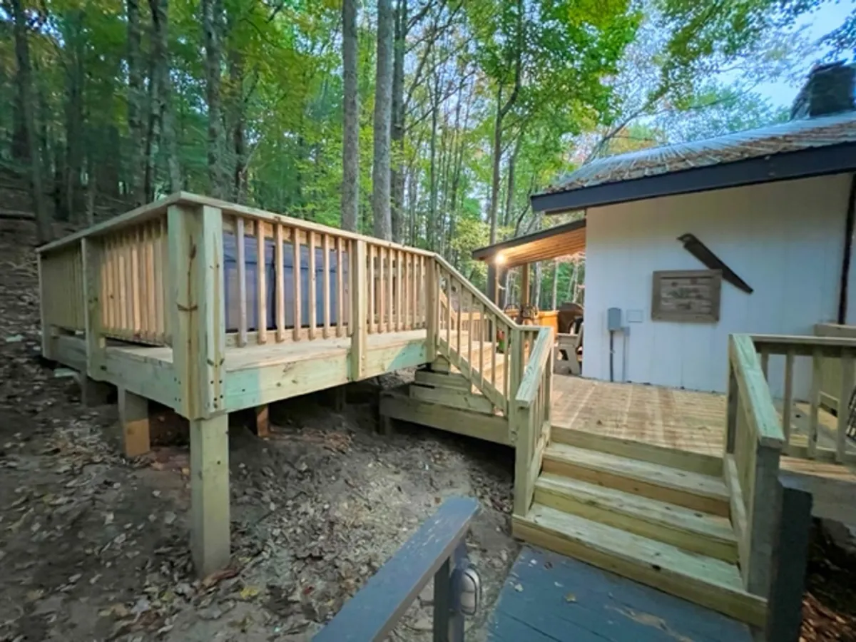 A wooden deck surrounded by mature trees, with steps leading up. The deck is spacious and situated amidst natural greenery, inviting relaxation and outdoor gatherings. The calm ambiance suggests it's suitable for all seasons, ideal for both sunny afternoons and crisp fall evenings.