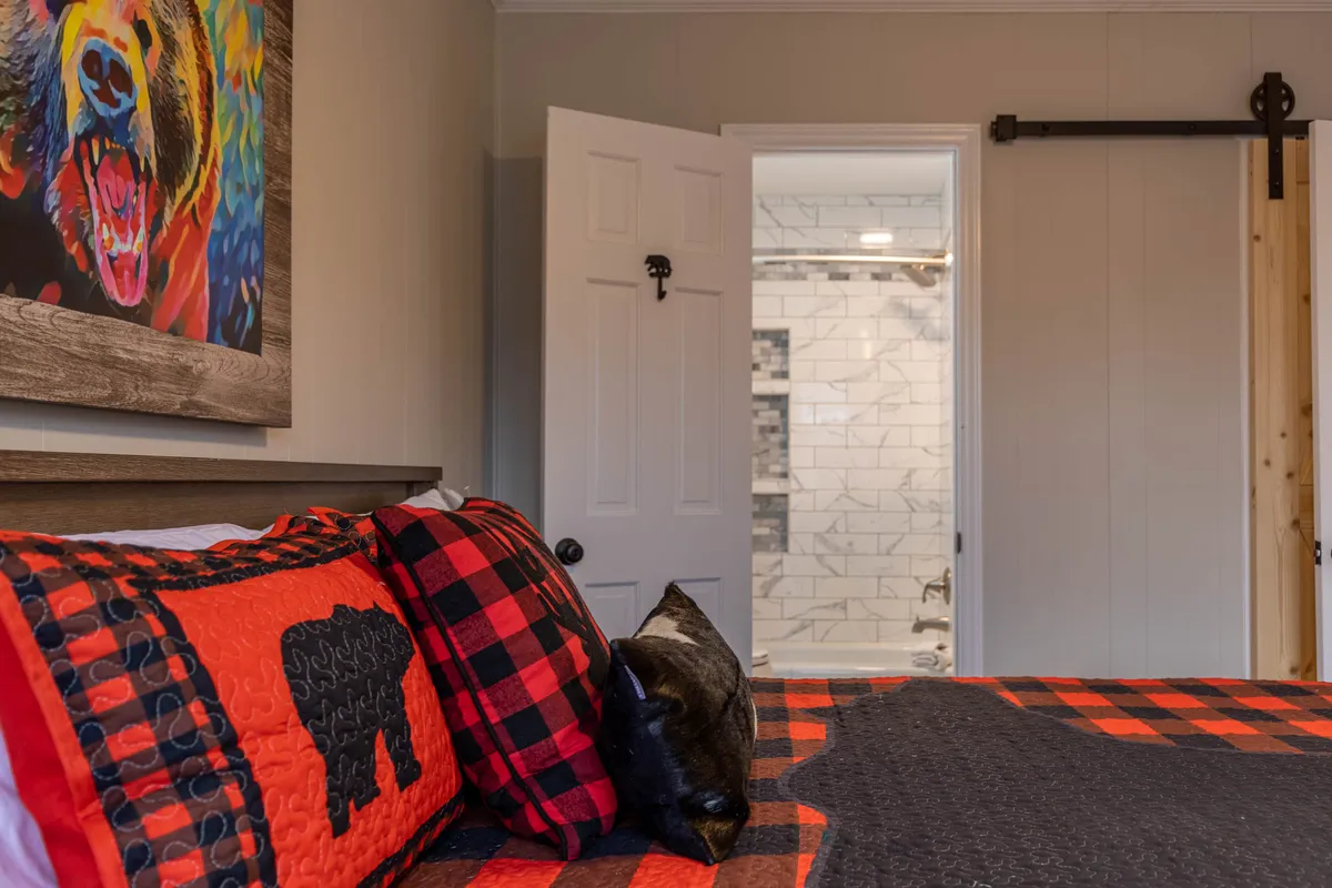 A welcoming bedroom with plaid bedding, vibrant bear artwork, and an open door leading into the bathroom. The rustic décor and soft lighting create a warm, inviting atmosphere perfect for relaxation and privacy.