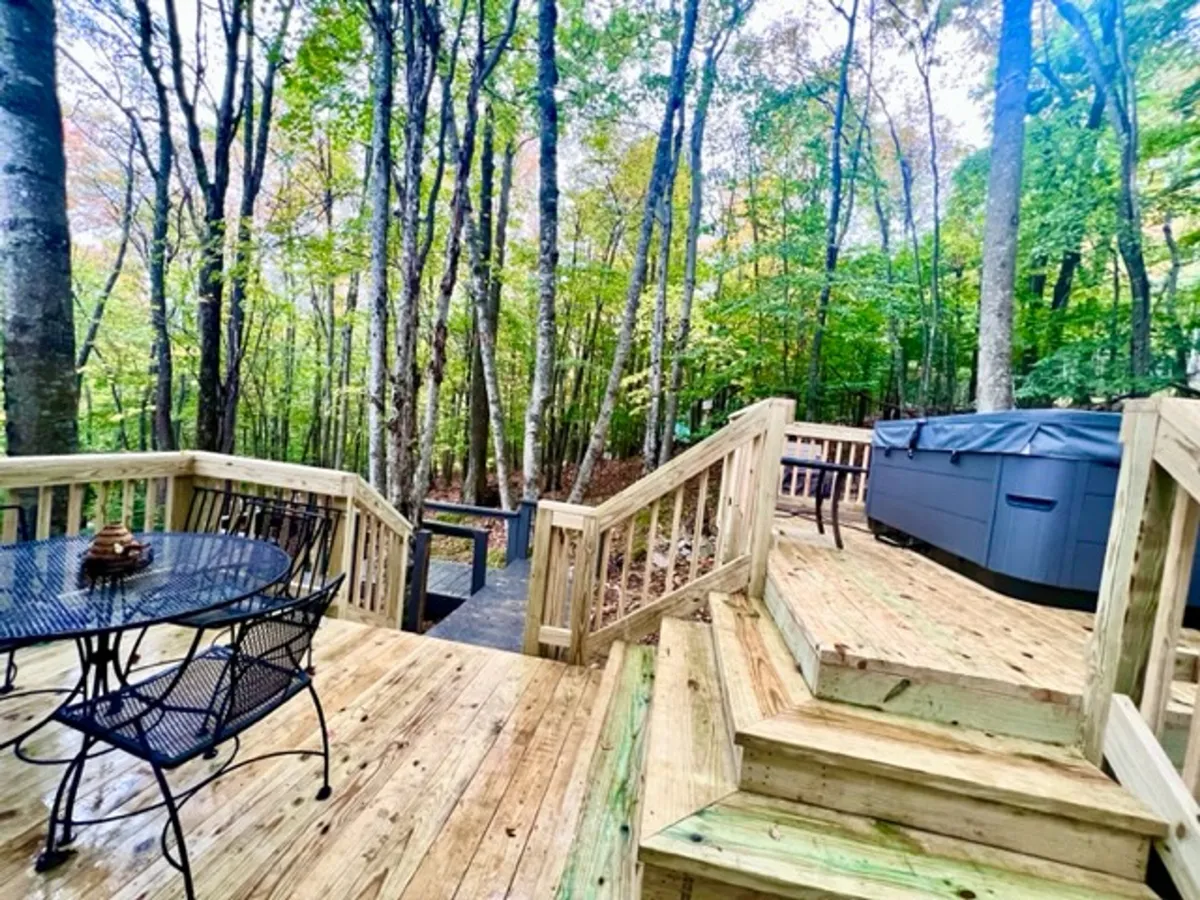 A spacious wooden deck is nestled among tall trees, featuring a large hot tub, an outdoor table with chairs, and the surrounding forest visible in the background. The scene suggests space for lounging in the hot tub, outdoor dining, and gatherings with friends—all while immersed in nature.