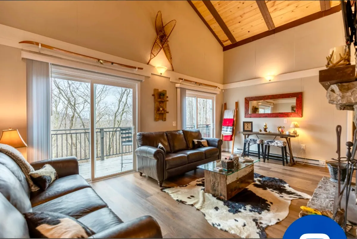 This interior image presents a spacious living area featuring a leather sofa, a cowhide rug, large windows with views of the outdoors, a wooden ceiling, and decorative ski equipment. The scene is welcoming and bathed in natural light, blending comfort with a distinctly mountain-lodge aesthetic.