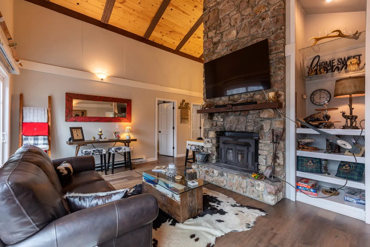 A warm and welcoming living room featuring a stone fireplace, leather sofa, wooden coffee table, and shelves adorned with personal decor. The inviting atmosphere is perfect for relaxing by the fire or gathering with friends and family, especially during colder months.