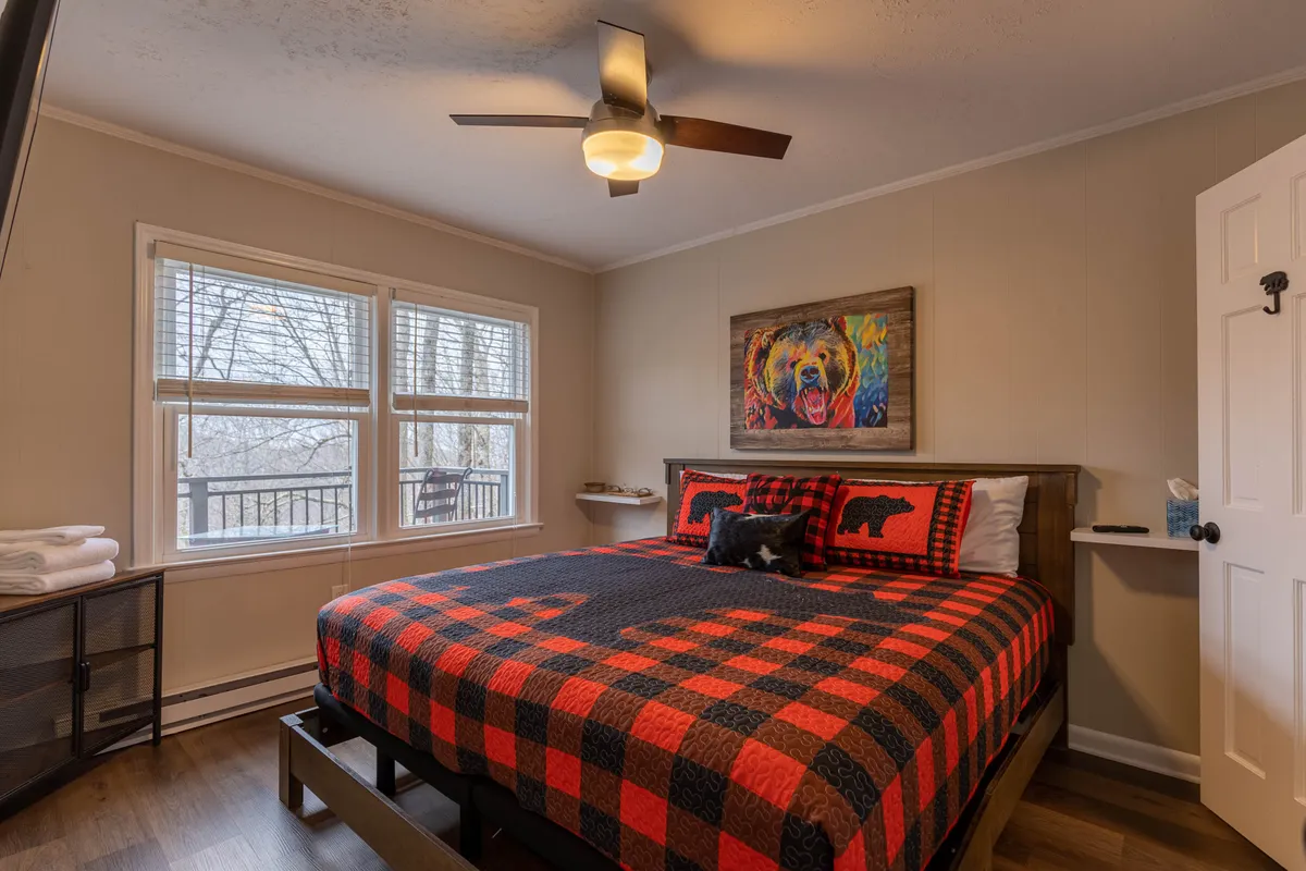 A cozy bedroom featuring a bed with a plaid blanket, rustic bear-themed artwork above the headboard, and large windows that let in abundant natural light while offering serene views of the surrounding woodland. The room exudes warmth and a cabin-style charm, emphasizing comfort and a connection to nature.