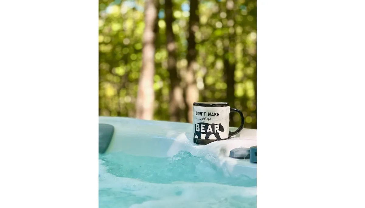 A tranquil hot tub overlooks a lush natural backdrop, with a coffee mug resting on the tub's edge. The scene evokes peaceful soaking, morning rituals, and a close connection to the wooded surroundings.