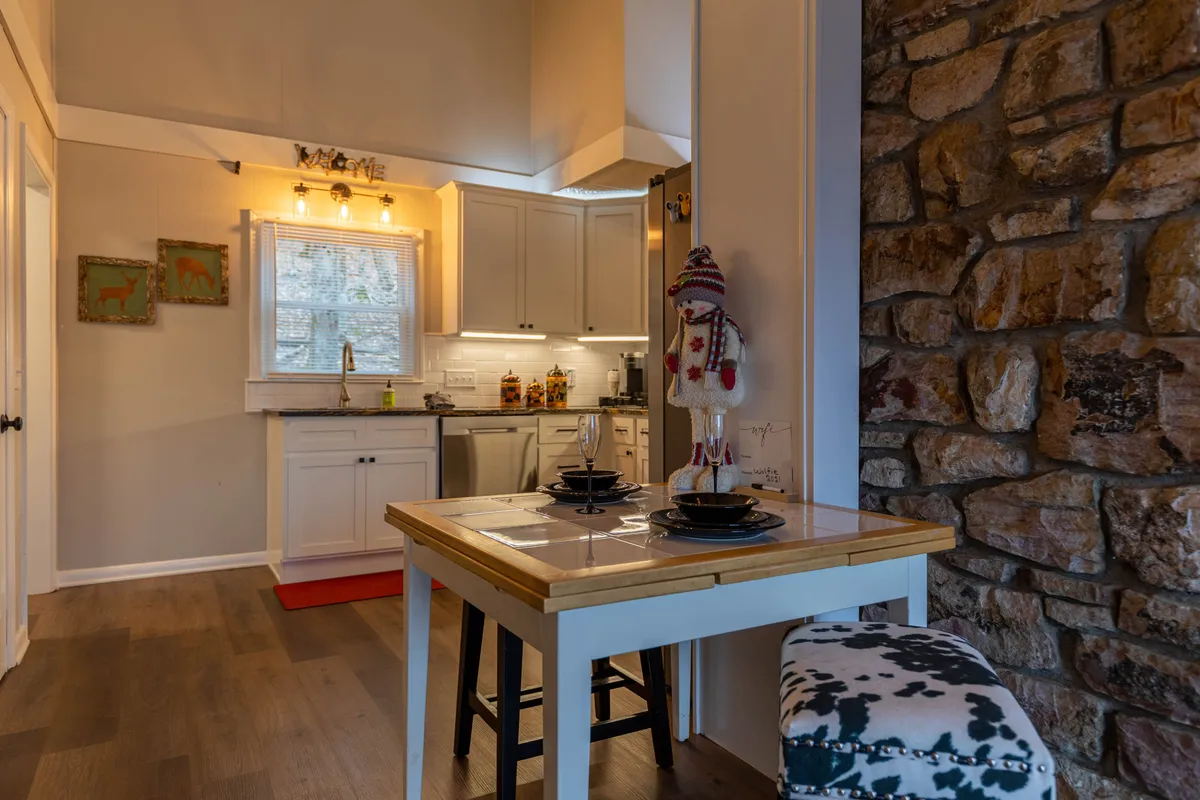 A view that captures the warm wooden dining table set beside a rustic stone wall, with the kitchen area visible in the background. Decorative touches, such as a playful snowman, enhance the welcoming atmosphere and highlight the easy flow between cooking and dining spaces.