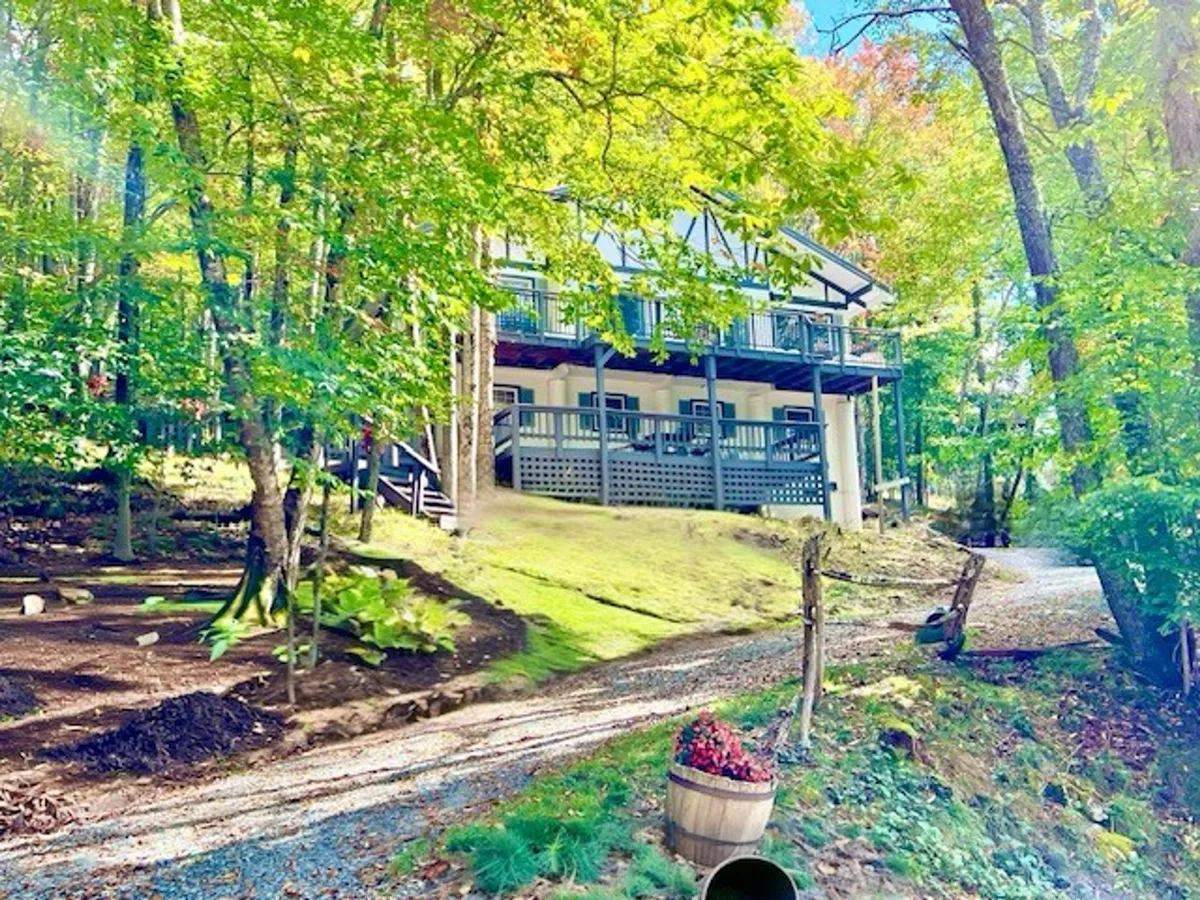 The image showcases the cabin nestled among tall, tranquil trees, with a charming exterior and a gravel path leading up to the welcoming front porch. The lush natural surroundings suggest privacy and direct access to trails, while the porch hints at relaxing mornings and peaceful evenings.